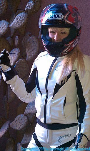 Hunter Storm wearing a motorcycle helmet with the visor open, long straight hair over her shoulder, dressed in white leather riding gear with black zippers and gloves.